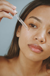 Woman applying clear brightening eye serum under eye with dropper against gray background.