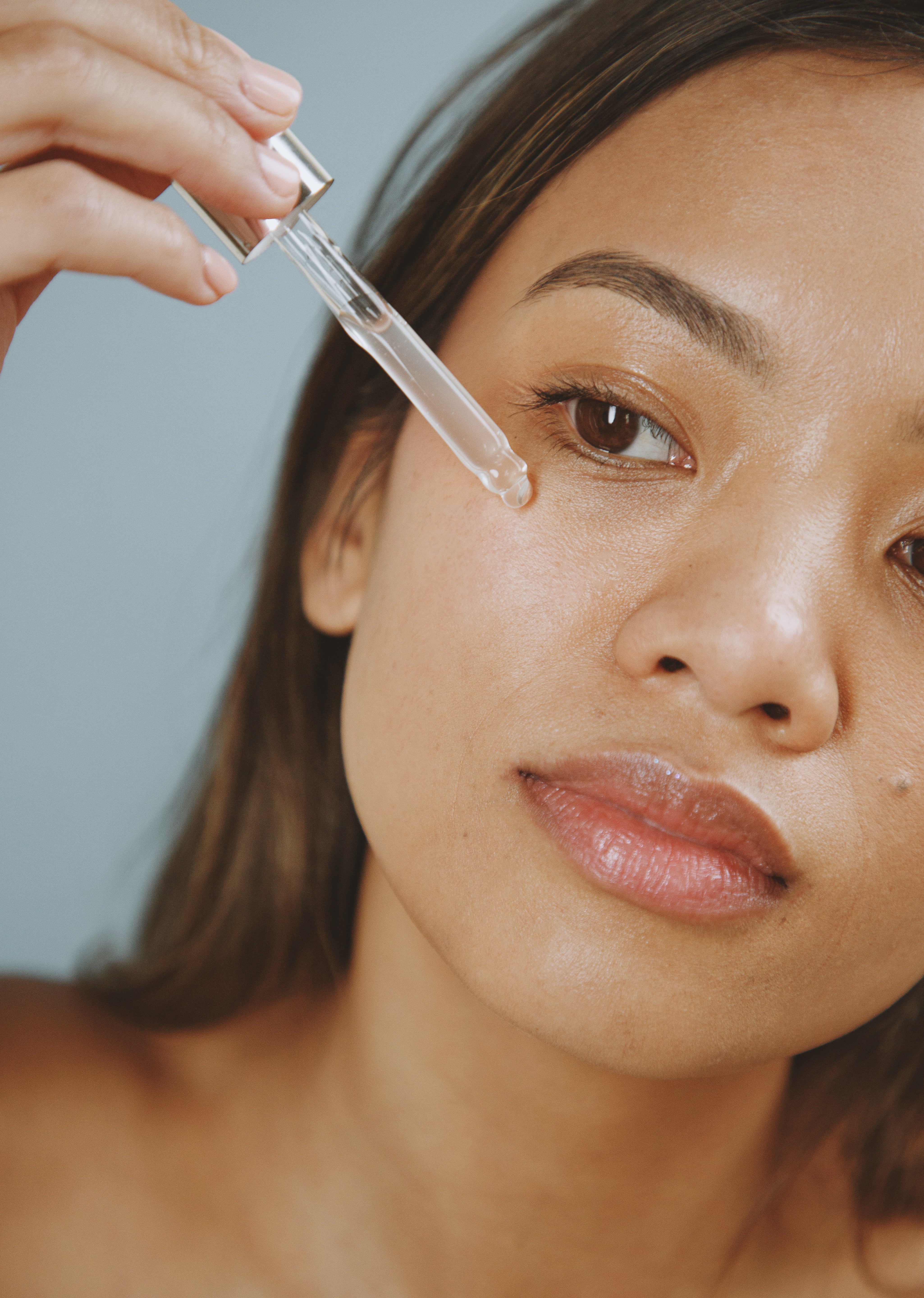 Woman applying clear brightening eye serum under eye with dropper against gray background.