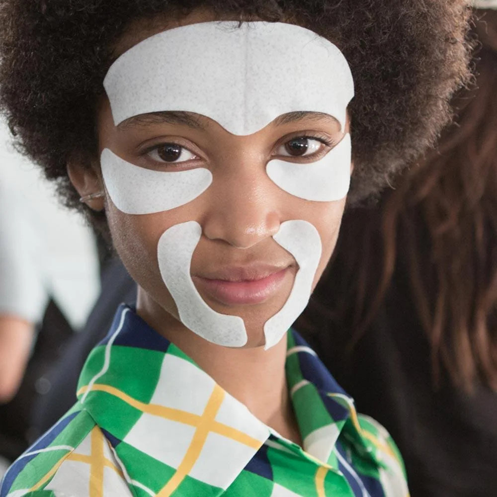 Woman wearing white face-lift lifting patches on forehead, cheeks, and chin with curly hair and green plaid shirt.
