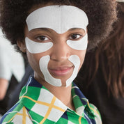 Woman wearing white face-lift lifting patches on forehead, cheeks, and chin with curly hair and green plaid shirt.