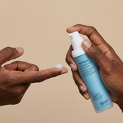 Matte blue cylindrical bottle with white pump dispenser held by brown-skinned hands, dispensing white lotion.